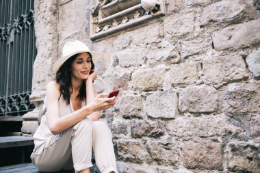 Genç bayan turist akıllı telefon kullanarak ve arkadaşından mesaj okuyarak şehrin yaz tatilinde taş binanın yanında otururken gülümsüyor.