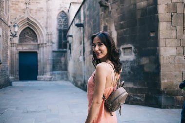 Şeftali rengi elbiseli, küçük modaya uygun sırt çantalı muhteşem yetişkin esmer kameraya bakıyor ve Gotik tarzda muhteşem mimarisi olan ortaçağ taş katedraline gülümsüyor.