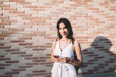 Beyaz elbiseli, sırt çantalı, elinde cep telefonu olan ve kameraya bakarken gülümseyen genç bir kadın.