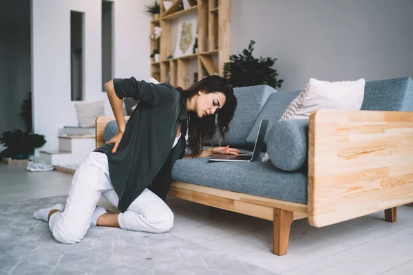 Stock image Young caucasian female suffer from back pain during remote job at home interior sitting near sofa, sad tired woman holding back feeling uncomfortable ache tension of spine arthritis chronic inflammation