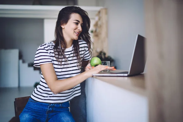 Cheerful young teen hipster girl spending free time at home searching information on web pages on laptop computer, positive caucasian woman freelancer enjoying remote job from home holding green apple