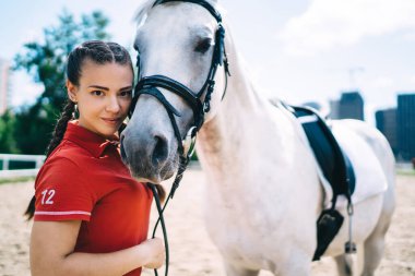Özel bir üniforma giymiş, beyaz şampiyon bir atla kameraya bakıp boş zamanların tadını çıkaran, binicilik sporu konsepti olan kaygısız genç bir kadının yarım boy portresi.
