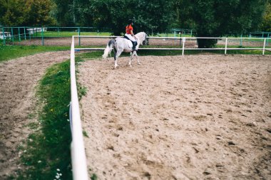 Siyah miğferli, kırmızı tişörtlü, çitin yanında beyaz ata binen çitli, yüzü olmayan kadın binicinin arka görüntüsü.