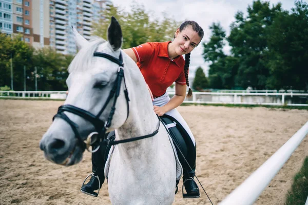 Female horse rider in red uniform standing near barrier on white horse during training on empty sand arena and looking  at camera