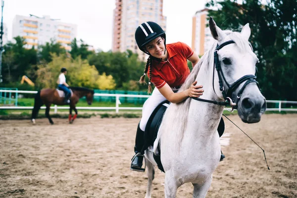 黒いヘルメットと赤いシャツの若い女性の馬術砂のパドックに鞍に座っていると白い馬の首を撫でる — ストック写真