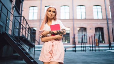 Eğitimli 20 'li yaşlardaki çekici bayan öğrenci ders kitabı ve akıllı telefon cihazıyla şehir caddesinde duruyor ve şöyle düşünüyor, güzel Y nesli cep telefonu şebekesi ile şehirde boş zaman geçirmek için.