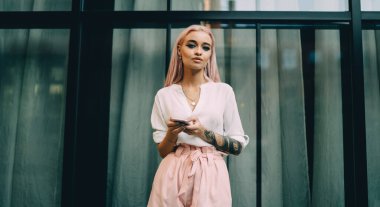 Çekici bir genç kızın yarım boy portresi. Dijital cep telefonu taşıyor ve şehir ortamında poz veriyor. Pembe saçlı ve akıllı telefonlu güzel bir kız kameraya bakıyor.