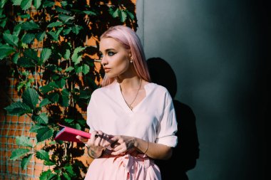 Zeki kız öğrenci, edebiyat kitabından gelen eğitim bilgilerini değerlendiriyor. Elinde not defteri tutan pembe saçlı, beyaz bir okul öğrencisi. Güneşli bir günde sokağın ortasına dikkatlice bakıyor.