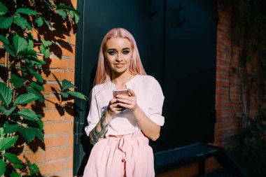 Güneşli şehirde boş zamanlarında kameraya gülümseyen renkli pembe saçlı mutlu bir kadın milenyumu portresi. Elinde akıllı telefon cihazıyla dışarıda poz veren güzel beyaz bir genç.