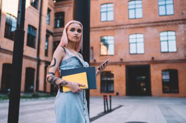 Çizim defteri ve not defteriyle dalgın dövmeli sanatçı şehir ortamında durup düşünceli hippi kız elinde dövmeli modern akıllı telefon teknolojisi olan blog yazarı.