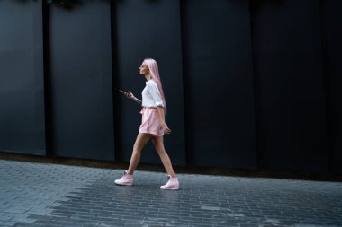 Elinde dijital akıllı telefonla gezen sıradan giyinmiş kadın turistin kentin tanıtım alanında yürümesi. Pembe saçlı hippi kız ellerinde cep telefonu teknolojisiyle şehri keşfediyor.