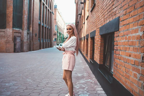 都会のストリートで笑顔を浮かべているケアフリー女性セルラーデバイスを介してウェブブログのための余暇時間を楽しんで 街を歩いている間にスマートフォンを使用してピンクの髪を持つ幸せな思春期の女性 — ストック写真