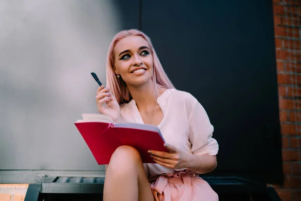 Cheerful female student with education planner resting at urbanity enjoying leisure time for creating interesting project ideas, happy contemplative schooler 20s holding textbook for learning