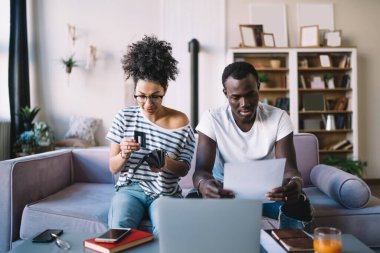 Afrikalı Amerikalı bir erkek evrak okurken, etnik bir kadın evde dizüstü bilgisayarla çevrimiçi ödeme yapmak için cüzdanından kredi kartını çıkarıyor.