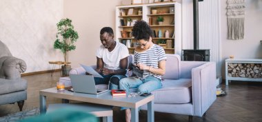 Çok ırklı genç bir çift kanepede rahatça kitap okuyor para sayıyor ve şömineli sıcak bir dairede ev faturalarını ödüyorlar.