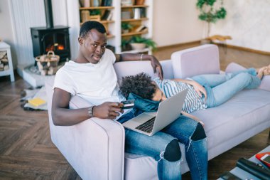 Afrika kökenli Amerikalı ciddi bir adam elinde kredi kartıyla dizüstü bilgisayarını açıp kameraya bakarken etnik bir kadın yakınlardaki kanepede uyuyor.