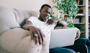 Beyaz tişörtlü yakışıklı, rahat koltukta rahatlayan, dizüstü bilgisayar kullanan, sırıtan ve evine bakan.