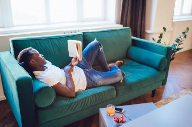 Tembel Afrikalı Amerikalı akıllı adam yalan söylüyor ve yakındaki kırmızı örtüde kitap okuyor. Akıllı telefon çileği ve meyve suyu ile. 