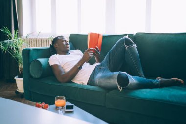 Afro-Amerikalı akıllı adam kırmızı kapaklı kitap okumaya ve akıllı telefon çileği ve meyve suyu ile yakındaki yeşil kanepede dinlenmeye odaklandı. 