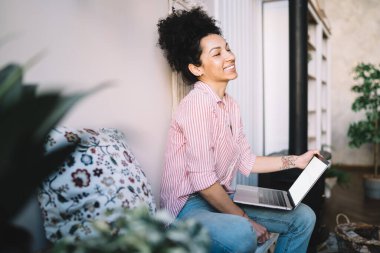 Evdeki kanepede bacak bacak bacak üstüne atarken dizüstü bilgisayar kullanan şık çizgili tişörtlü sakin etnik kadın. 