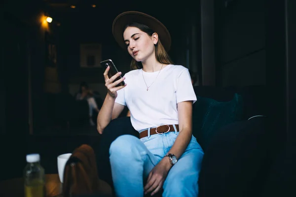 流行の帽子とカジュアルな服装で若い肯定的なスタイリッシュな女性は 現代のカフェテリアでアームチェアに座って スマートフォンを使用して友人とチャット — ストック写真
