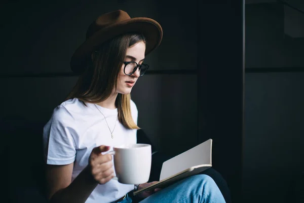 坐在扶手椅上 手里拿着一杯热饮时 头戴时髦帽子 戴着眼镜 沉思时尚的年轻女士在抄本上读笔记的侧视图 — 图库照片