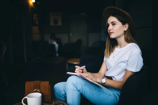 穿着时髦帽子和休闲装的年轻时髦女士坐在咖啡店的扶手椅上 一边看着一边记笔记的侧影 — 图库照片