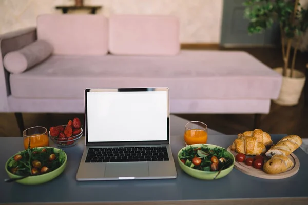 Opened laptop with blank screen next to healthy salad and pastry with juice on table in modern living-room by couch in daylight