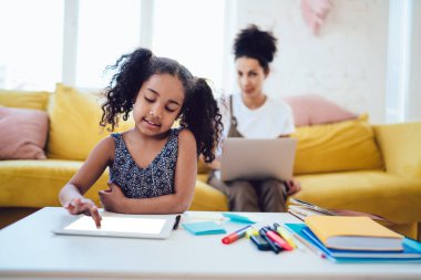 Meraklı siyah kız masanın önünde oturuyor ve boş tablet ekranına dokunuyor. Annesi de dizüstü bilgisayarla çocuğa bakıyor.