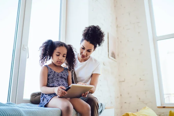 穿着蓝色衣服的非洲裔美国女儿低角度坐在窗台上 在家里给妈妈展示着白色衬衫平板电脑 — 图库照片