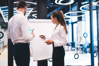 Beyaz tahtanın yanında duran ve modern ofiste projenin detaylarını tartışan şık takım elbiseli başarılı iş arkadaşlarının arka planı