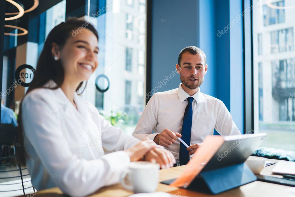 Smiling male and female coworkers in formal wear talking with pleasure about business using tablet while sitting at table in well lit office