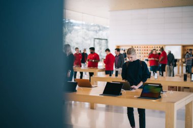 Malları incelerken masanın yanında duran genç bayan müşteri, mağaza çalışanları ve ziyaretçiler arka planda bulanık duruyorlar. Milenyum yeniyetme kız satın almak için aygıtı kontrol ediyor