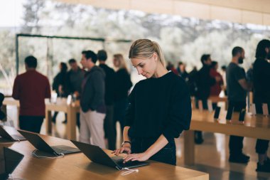 Malları incelerken masanın yanında duran genç bayan müşteri, mağaza çalışanları ve ziyaretçiler arka planda bulanık duruyorlar. Milenyum yeniyetme kız satın almak için aygıtı kontrol ediyor