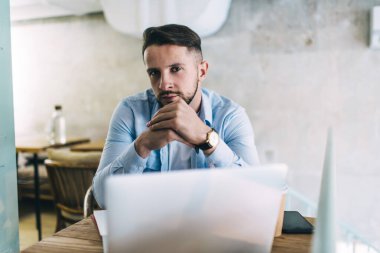 Sakallı, kara kara düşünen, laptoplu cep telefonu ve kafeteryada bir fincan sıcak içecekle masada oturan yakışıklı bir erkek.