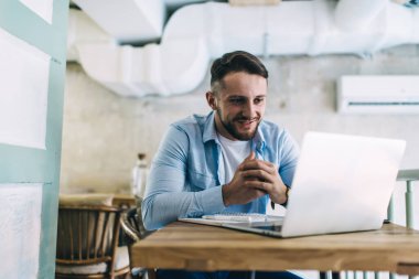 İyi aydınlatılmış kafedeki ahşap masada oturmuş dizüstü bilgisayara bakan rahat ve yakışıklı bir erkek.