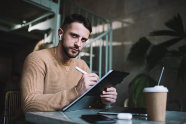 Kablosuz kulaklık takan, kafede oturmuş kameraya bakarken notlar yazan sıradan giyinmiş genç bir erkek.