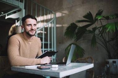Kahvede elinde dizüstü bilgisayarla, elinde bej renkli bir süveterle oturmuş gülümseyen ve başını çeviren neşeli genç bir erkek.