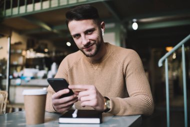 Kafede akıllı telefon kullanarak otururken kablosuz kulaklık takmış, hafifçe gülümseyen ve kameraya bakan genç sakallı bir erkek.