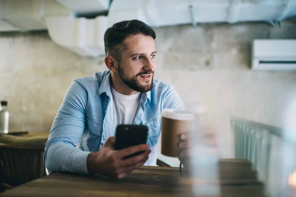 턱수염을 긍정적 남자가 뜨거운 커피를 마시고 탁자에 고개를 돌리는 스마트폰을 — 스톡 사진