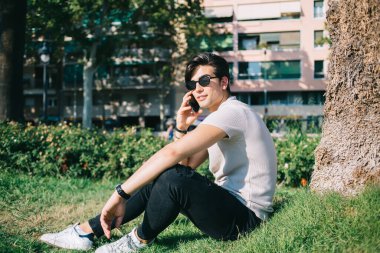 Havalı giyinmiş beyaz adam bahar tatillerinde şehir parkında oturuyor cep telefonuyla konuşuyor yakışıklı erkek turist uluslararası bağlantıyı akıllı telefon görüşmesi yapmak için kullanıyor.