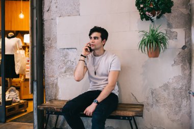 Düşünceli etnik erkek, gündelik giysiler giyip Barselona 'da taş binanın yanındaki ahşap bankta oturuyor ve rahatlarken ve başka tarafa bakarken cep telefonuyla konuşuyor.