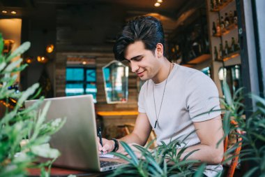 Başarılı düşünceli erkek yazar serbest yazar blog yayını için makaleler hazırlamaya odaklanmış, online proje için çizimler yaparken gülümseyen genç adam kafeteryada dizüstü bilgisayarla oturuyor.