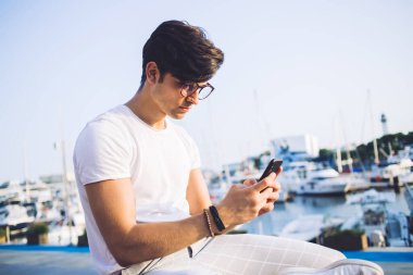 Kafkasyalı beyaz adam blogcu uygulama ve 4G bağlantısını kullanarak cep telefonundan e-posta yolluyor. Gelir mesajlarını kontrol eden ciddi bir erkek gezgin tatillerde liman yakınlarında dinleniyor.