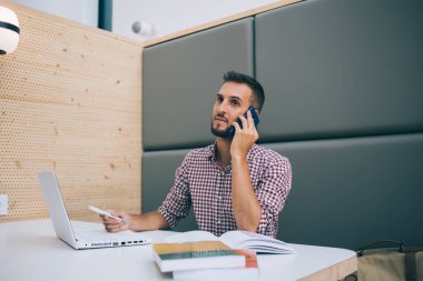 Modern işyerindeki masada oturan meslektaşımla cep telefonuyla konuşmak için dizüstü bilgisayar kullanan gündelik kıyafetlerle etnik ve ciddi bir serbest çalışan.