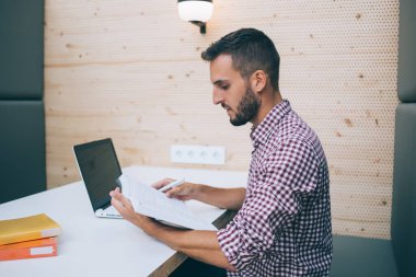 Gündelik giysiler içinde, bilgisayarla belgelere bakıp modern işyerindeki masada oturan serbest çalışma projesiyle meşgul etnik bir işçi.