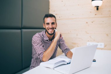 Heyecanlı etnik gülen serbest yazar elinde kalemle dizüstü bilgisayar kullanarak modern kafede oturup kameraya bakarak yeni bir başlangıç yaratıyor.