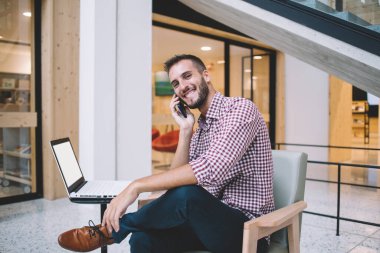 Keyifli gülümseyen, sakallı, normal tişörtlü, cep telefonuyla konuşan alışveriş merkezinde dizüstü bilgisayarlı genç bir adam.