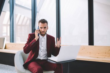 Resmi olarak asık suratlı bir etnik erkek, partneriyle iletişim kurarken modern iş yerindeki masada otururken akıllı telefon hareketleriyle hayal kırıklığına uğrar. 