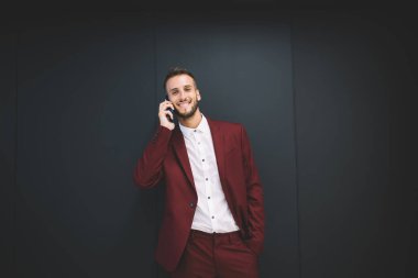 Beyaz gömlekli, siyah düğmeli, kırmızı ceketli ve pantolonlu neşeli genç sakallı adam gri arka planda cep telefonundan arıyor.